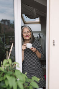 Portrait of happy senior woman leaning at opened balcony door with sup of coffee looking at distance