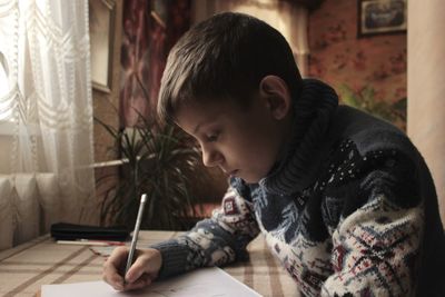 A boy in a christmas sweater sits at the table and draws and writes