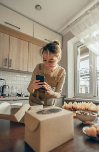 Happy blogger bakery use smartphone, standing by take out delivery box for customer order in kitchen