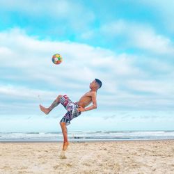 Full length of shirtless man playing soccer at beach against sky