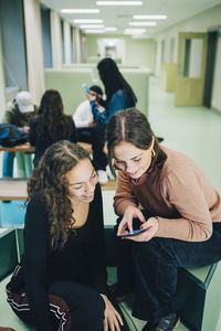 High angle view of teenage girl showing smart phone to female classmate while sitting at corridor in school