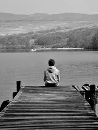 Rear view of boy sitting on pier over lake against sky