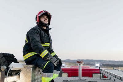 Pensive adult male wearing protective hardhat and protective suit while sitting on top of fire engine and looking away with clear sky on background