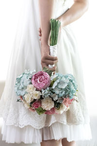 Midsection of bride holding bouquet while standing against white background