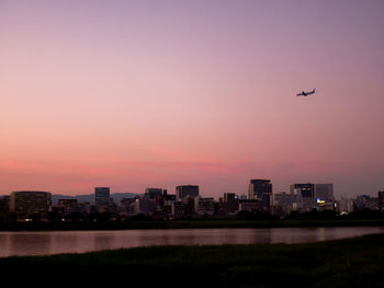 Airplane over river against sky in city during sunset