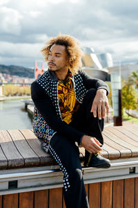 Young bearded african american hipster guy with curly afro hair dressed in stylish colorful shirt and black jeans looking away while sitting on border against blurred urban background