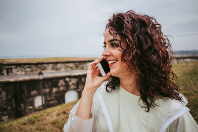 Smiling woman talking over phone while standing on grass against sky