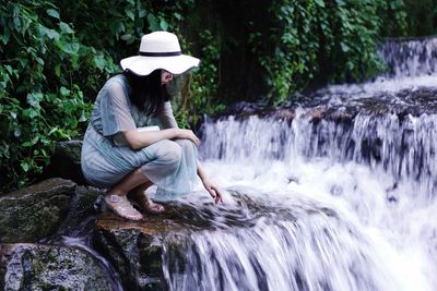 Full length of woman sitting on rock by waterfall