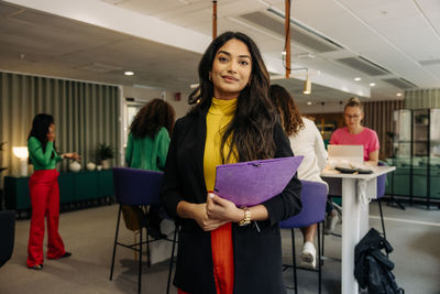 Portrait of smiling businesswoman holding folder while standing at workplace