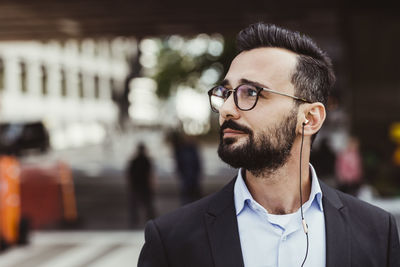 Confident businessman with in-ear headphones looking away while standing outdoors