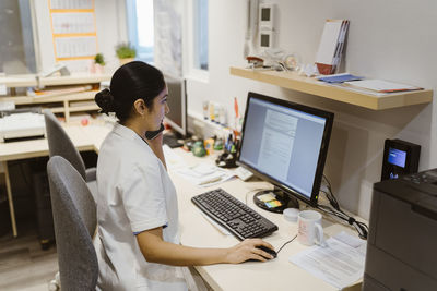 Female healthcare worker using computer while talking on smart phone at clinic