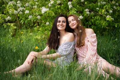 Two young girls in dresses are sitting on the green grass under a white tree and laughing