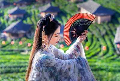 Beautiful woman in traditional clothing holding hand fan sitting at field
