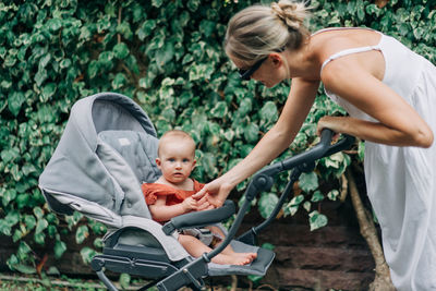 Woman in sundress talking calmly to infant while pushing stroller along sidewalk on green street