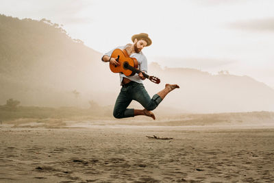 Full length of man jumping while playing guitar at beach