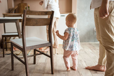 Little baby walks by wooden floor in kitchen between parents legs.