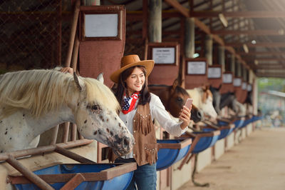 Smiling young woman taking selfie with horse while standing in stable