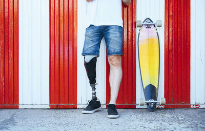 Young man with leg prosthesis standing next to skateboard at a wall