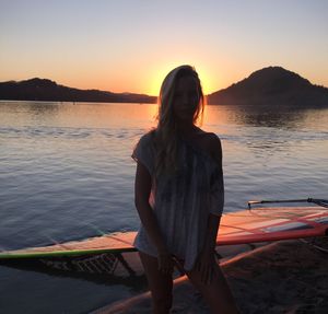 Young woman standing by windsurfing board at lake against sky during sunset