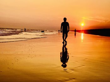 Rear view of silhouette man walking on shore at beach against sky during sunset