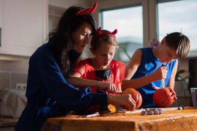 Mother, daughter and son in halloween costumes carve eyes and teeth on pumpkins in a darkened room