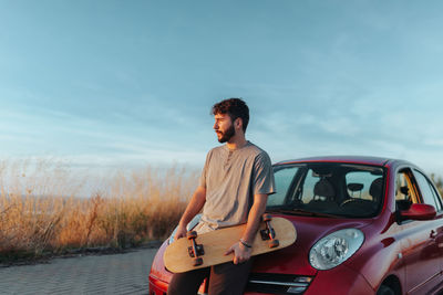Young man looking away while standing on car against sky