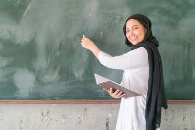 Portrait of teacher wearing traditional clothing writing on blackboard while standing in classroom