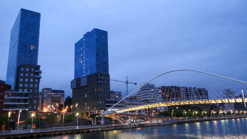 Illuminated bridge over river by buildings against sky in city