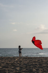 Boy flying kite while standing at beach against sky during sunset