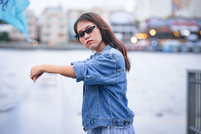 Beautiful young woman standing by railing against river and city in background