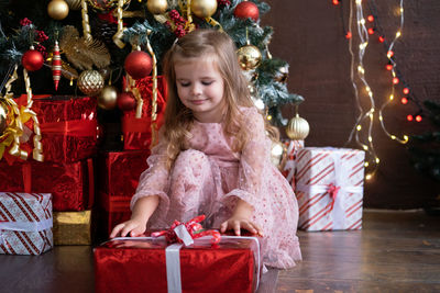 Happy little smiling blond girl in beautiful dress with gift box near christmas tree