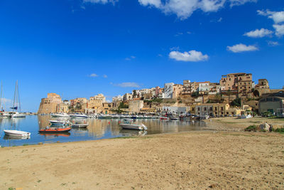 Sailboats moored on sea by buildings against blue sky