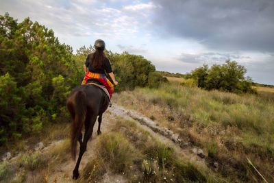 Rear view of woman riding horse on field against cloudy sky