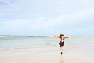 Rear view of woman with arms outstretched walking at beach against sky