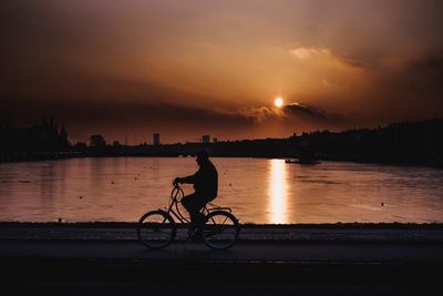 Silhouette man with bicycle on river against sky at sunset