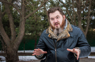 Confused bearded man wearing jacket against trees during winter at park