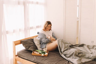 A cheerful young teenage woman plays with her pet a small dog in bed in the morning in a cozy house