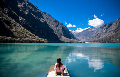 Rear view of woman sitting on pier at lake against mountains