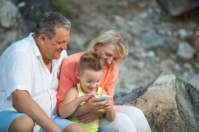 Smiling grandparents sitting with grandson using mobile phone at beach