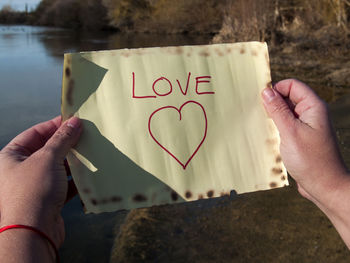 Cropped hand of woman holding text and heart shape on paper against lake