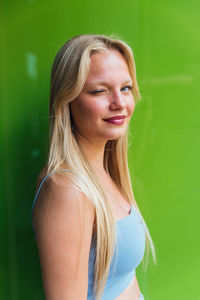 Side view of coquettish female with blond hair winking at camera on green background