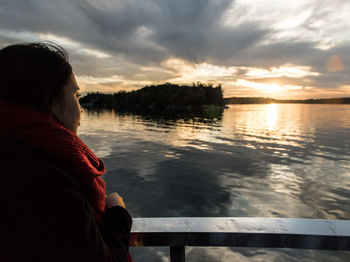 Rear view of woman sitting by lake against sky during sunset