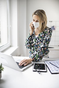 Businesswoman wearing mask using laptop while talking on phone at home