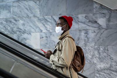 Pensive african american millennial man with smartphone on escalator subway wearing protective mask