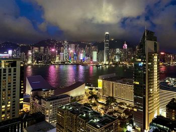Illuminated modern buildings by river against sky at night