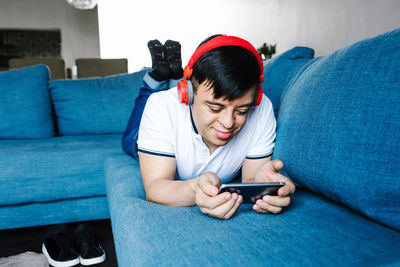 Delighted focus latin teen boy in headphones playing videogame on mobile phone while lying on sofa at home