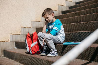 Full length of boy sitting on steps with backpack during sunny day