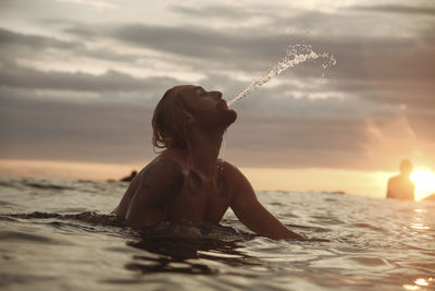 Silhouette man spraying water while surfing on sea against sky during sunset