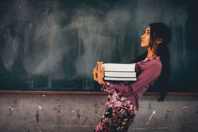 Side view of young woman with books looking up while standing by blackboard in classroom