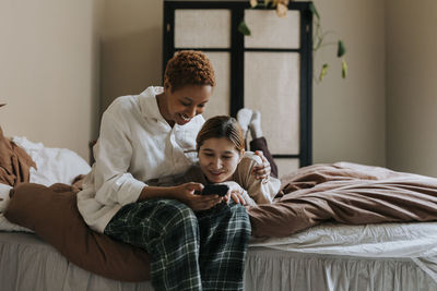 Smiling friend sharing smart phone with non-binary person lying on bed at home